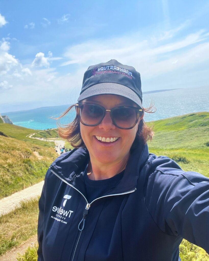 Smiling woman in sunglasses, hat, and jacket, taking a selfie with ocean, cliffs, and path in the background.