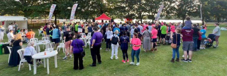 Corporate festival: large group of people gathering outdoors on grass. Tents and flags in background.